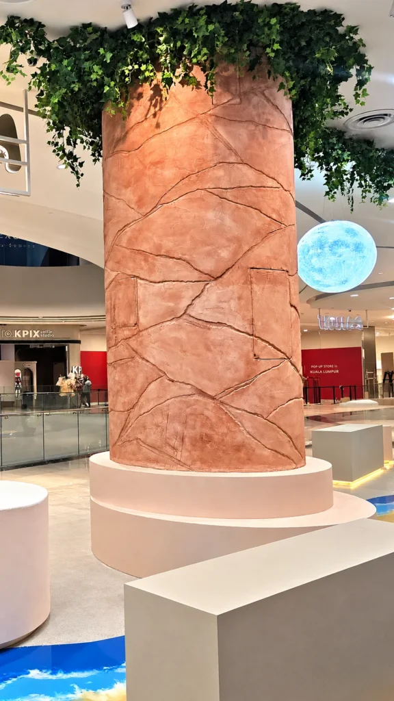 Full-height view of the terracotta-toned StoneArt column at Ombak KLCC, with foliage above and a pale tiered base.
