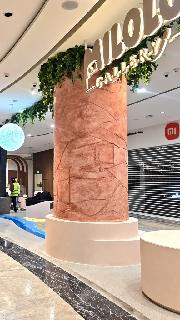 Wider view of the StoneArt feature column at Ombak KLCC with overhead signage, a pale base and blue floor detailing.
