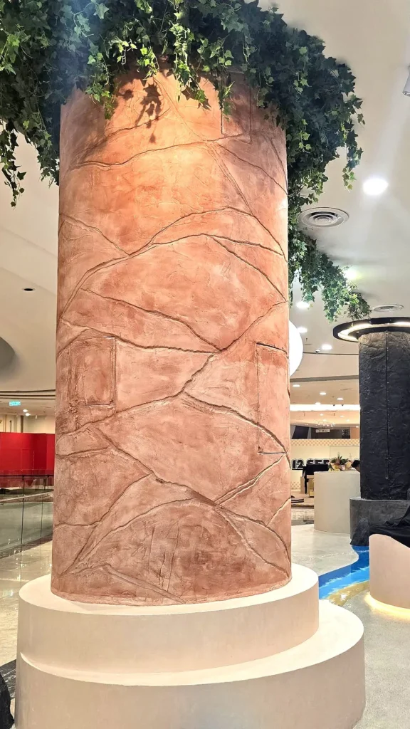 StoneArt column at Ombak KLCC showing irregular recessed lines and warm terracotta tones beneath green foliage.